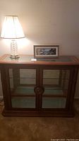 Front view of wood and glass display cabinet with lamp and framed print on top, placed against a wall