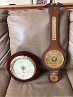 Two barometers pictured on a beige couch: one round RSVP barometer and one larger Baromaster barometer with thermometer and hygrometer.