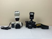Front view showing Minolta film camera with flash and lens next to Nikon digital camera with flash and strap
