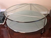 Two-tier round coffee table with clear glass top, frosted glass bottom, metal frame, and casters. Visible triangular metal support under the top glass.