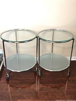 Front view of two round glass end tables with metal frames and casters on wood floor against wall.