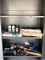 Painting supplies arranged on metal shelves including spray paint cans, plastic sheet, drop cloths, paint rollers, brushes and trays.