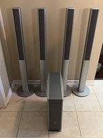 Four silver tall tower speakers with round bases standing upright on a tiled floor alongside the subwoofer.
