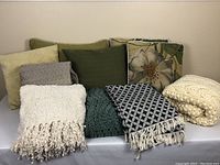 Photo shows six throw pillows in yellow, green solid tones and floral pattern, arranged with five blankets in various textures and colors including cream knit, gray quilted, white fringed, dark green knit, and black and white patterned.