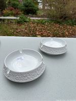 Two white bisque porcelain Royal Bavaria ashtrays shown together on outdoor surface, textured sides visible, one slightly raised behind the other.