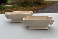 Two creamware rectangular planters with wide angular handles and pedestal bases shown on white surface outdoors with fall foliage. Visible crazing and dirt marks on surface.