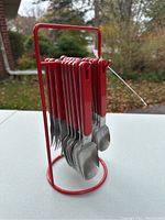 Cutlery set with red handles and red metal stand, showing full front view of knives, forks, and spoons hanging on stand.