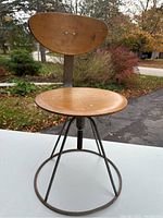 Front view of small chair showing round wooden seat and rounded triangular wooden backrest fixed with screws. Metal frame forms circular base with three metal legs supporting seat.