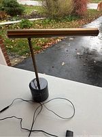 Full view of desk lamp showing brass-tone cylindrical light bar, vertical rod, black marble base and attached cord with plug on white surface.
