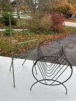 Two metal magazine racks on an outdoor table, one green tripod style and one black curved rack