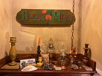 Wide view of the lot showing oil lamps, various wooden candlesticks, small decorative figurines, and the wooden sign above