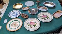 Photo showing assorted decorative and serving plates and dishes on green tablecloth, including floral, commemorative, brass and silver items.