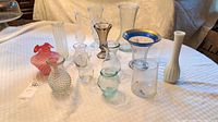 View of all 11 vases on a checkered tablecloth including varied shapes, sizes, and colors such as red-pink glass, clear glass, blue rimmed, aqua tinted, and white ceramic