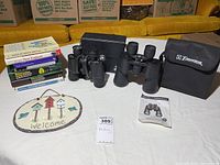 Overall lot view showing birding books stacked, two pairs of binoculars, black binocular case, and decorative welcome sign with birdhouses and birds design.