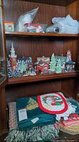 Photo showing Department 56 Christmas village buildings, trees, and figurines arranged on a shelf with holiday linens below.