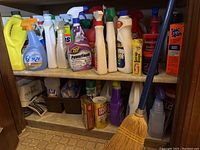 Wide view of multiple cleaning supplies including spray bottles, liquid containers, and a broom inside a cupboard.