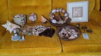 Full view of various coastal decor items including large conch shell, ceramic bowl filled with seashells, shell wreath, two lighthouse figurines, glass containers with shells, and metal wall hooks placed on yellow sofa.