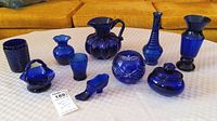 Full lot display showing ten cobalt blue glass pieces on a white tablecloth, including a pitcher, basket, vases, container, decorative shoe, and small bowls.
