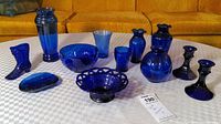 Overview of eleven cobalt blue glass pieces arranged on table including bowls, vases, glasses, soap dish, candlesticks, and boot-shaped piece