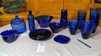 Photo showing full assortment of cobalt blue glass items on table including glasses, bowls, plates, jar with lid, bottle, cup, and vase.