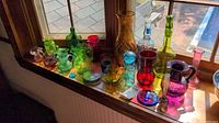 Wide view of colorful glass bottles, pitchers, vases arranged on windowsill in sunlight showing various shapes and colors including green, pink, red, and blue.