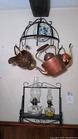 Full view of two copper kettles, copper watering can, ceramic bowl in hanging metal cage rack, and wall mount rack holding two oil lamps below.