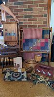Photo showing small wooden wall cabinet with painted village scene and wooden quilt rack holding two quilts, plus woven baskets and other country decor items on carpeted floor.