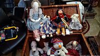 Photo showing multiple rag dolls and stuffed animals inside and on top of a wicker trunk, illustrating the lot's composition and condition.