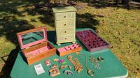 Three jewelry storage boxes with assorted costume jewelry displayed on green outdoor table, shown in daylight