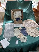 Full view of assorted lace and crochet items including box with small crochet pieces and several large doilies and table runners laid out on green fabric.