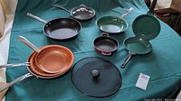 Overall view of all frying pans and spatter guard arranged on green cloth, showing copper and black pans plus green double-sided pan and small pan.