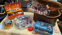Wide view of the sewing materials including orange and blue sewing boxes, clear round spool container, tin with buttons, and tomato pin cushion.