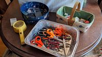 Photo shows three containers: a blue floral fabric-covered box containing scissors and sewing tools, a foil tray holding approximately 20 pairs of scissors with orange, black, and metal handles, and a light green and white basket containing sewing notions and thread spools. Also two yellow handheld magnifying glasses.