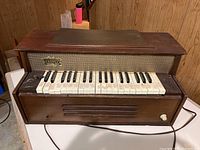 Front view of vintage Emenee electric pipe organ showing wooden body, 32-key keyboard, control knob and Emenee badge on speaker grille.