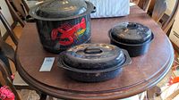 Black and white speckled enamelware set on wooden table including lobster pot and two roasting pans with lids.