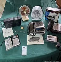 Multiple vintage electronics items laid out on a green cloth including Sony Watchman, clock radios, fans, and accessories.