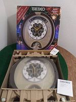 Clock in original protective packaging with plastic cover and mounting hardware visible.