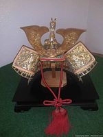 Front view of miniature kabuto helmet on wooden stand with black lacquer platform, gold-tone metal crest and red tassel cord
