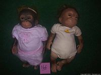 Two baby monkey dolls placed side by side on a green surface; one wears a pink ballerina dress and the other a white onesie.