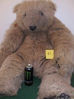Front view of the large beige plush teddy bear sitting upright with a Monster energy drink can at its feet for size scale. The bear has long limbs and a sewn black nose with black eyes. A small yellow note "41" is attached to the bear's chest.