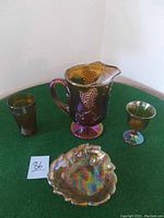 Complete lot showing a pitcher with handle and grape cluster pattern, two different glasses, and a decorative leaf-shaped bowl, all on a green tablecloth.