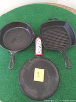 Three cast iron pans on green felt, showing different shapes and surfaces including a grill pan, skillet, and round pan.