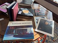 Wide shot of all coffee table books including Edward Hopper art book, three American Heritage history books, atlas, and several other books and prints scattered on wood table near window.