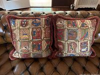 Two square throw pillows side by side on a brown tufted leather surface, showing the medieval-style printed side with rust-red textured fringe.