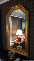 Front view of tall gold wood framed mirror with clear glass jar with a lid on black surface below.