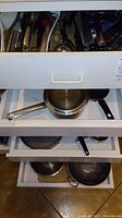 Four open kitchen drawers showing pots, pans, lids, and cooking utensils including strainers and frying pans.
