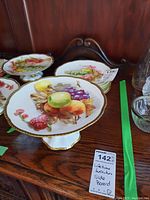 Meissen cake stand and six fruit plates with detailed hand-painted fruits and gold trim, displayed on a wood sideboard.