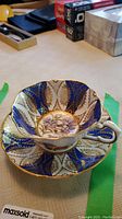 Side view of porcelain teacup and saucer with blue and gold trim, displaying floral cornflower design in the center.