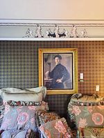 Wide view of the framed print on a patterned wall above floral cushions, showing the full ornate gold frame and wallpaper context