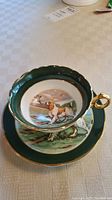 Top-down view of teacup showing hunting dog painting inside, green border, and gold handle.